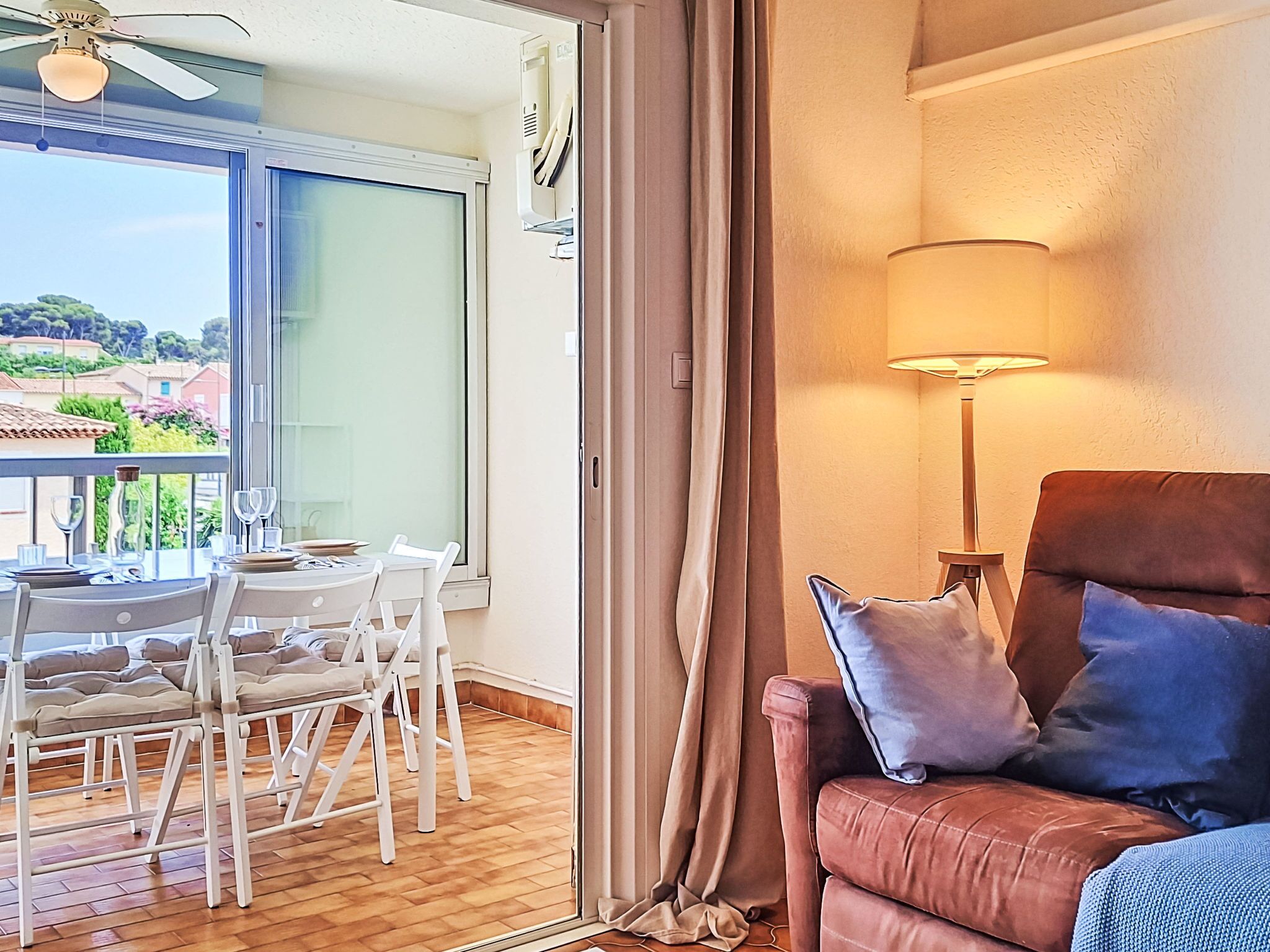 A room with a dining table set for two, a brown leather sofa with cushions, a standing lamp, and a view of a balcony with a table and chairs.