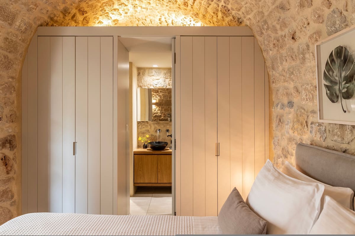 Un dormitorio con cama, mesita de noche, ventana grande y pared de piedra.