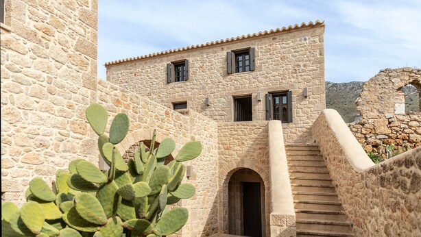 A stone building with a staircase leading to an entrance, a cactus plant in the foreground, and a clear sky.