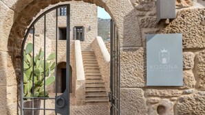 A stone building with a sign for 'Korona Boutique Hotel' and a cactus plant visible through a metal gate.