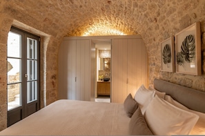 A bedroom with stone walls, a bed with pillows, a wardrobe, and a framed plant.