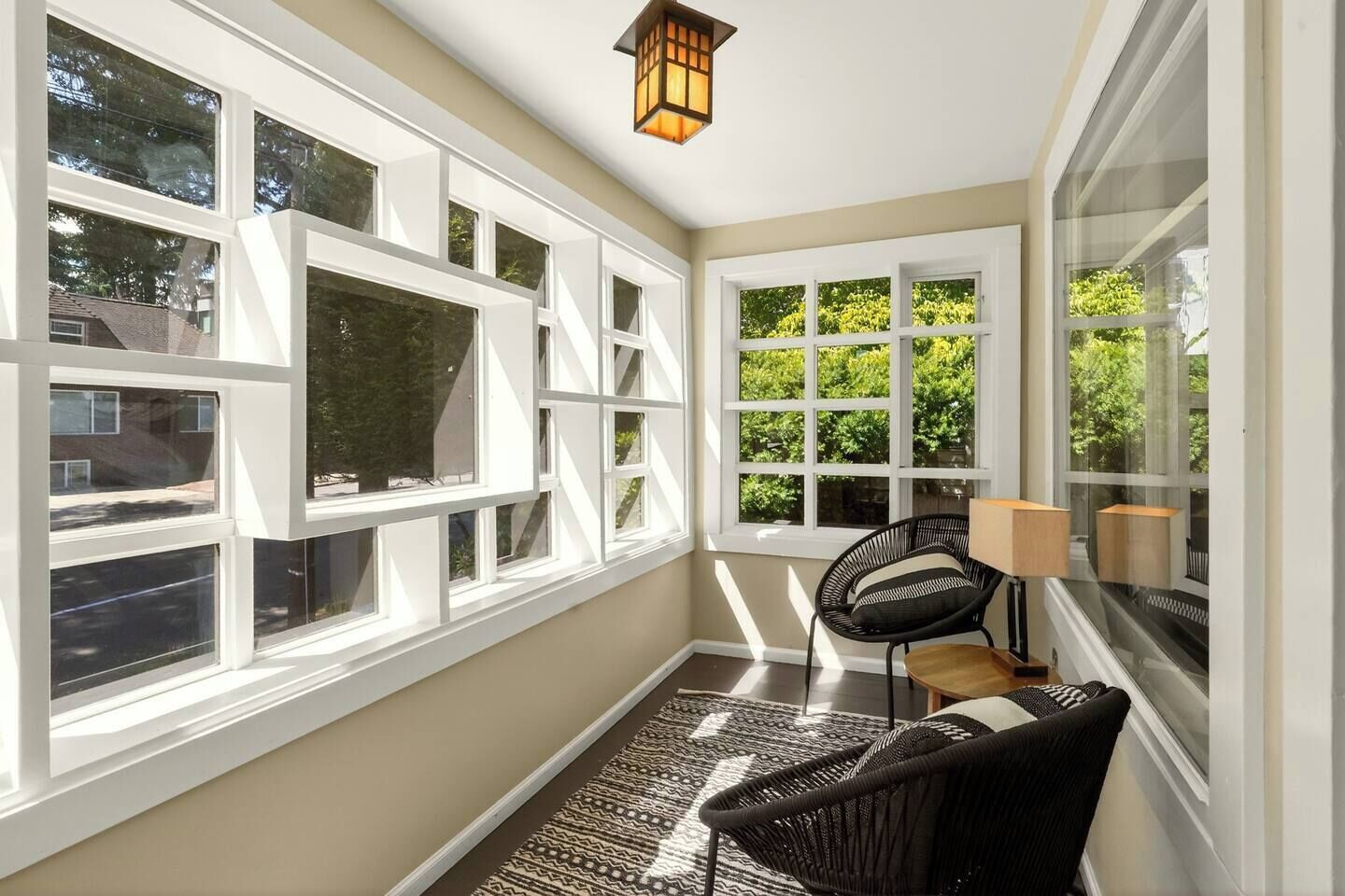 Una habitación soleada con ventanas amplias, una silla de mimbre negro, una pequeña mesa y una lámpara de pie.