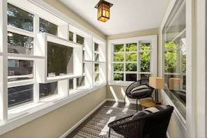 A sunlit room with large windows, a black wicker chair, a small table, and a floor lamp.