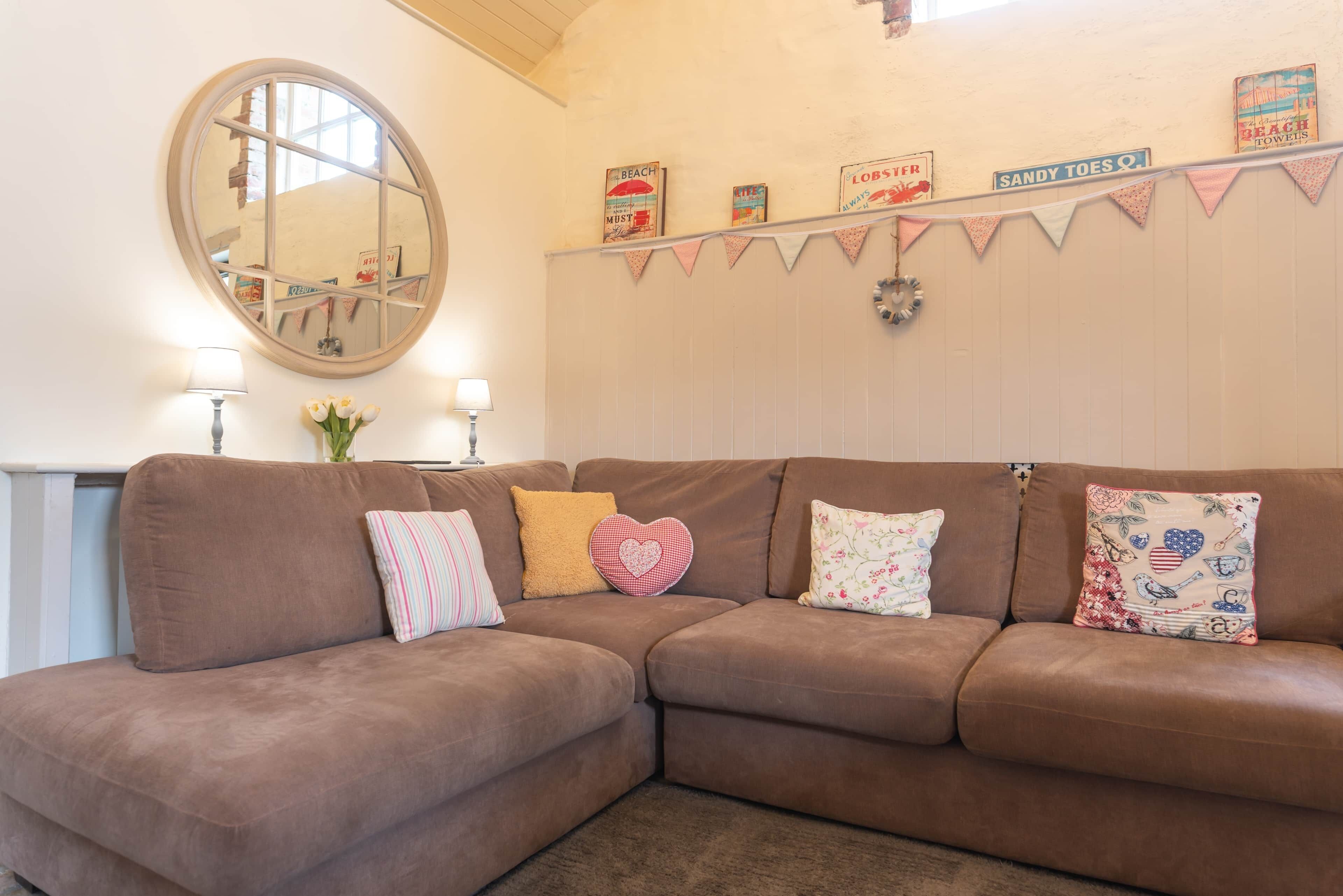 Ein braunes Ecksofa mit herzförmigen und floralen Kissen, einem runden Spiegel sowie Wanddekorationen, darunter ein Strandschild und Wimpelketten.