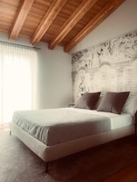 Un dormitorio con cama, vigas de madera en el techo, un mural en la pared y una ventana con cortinas.