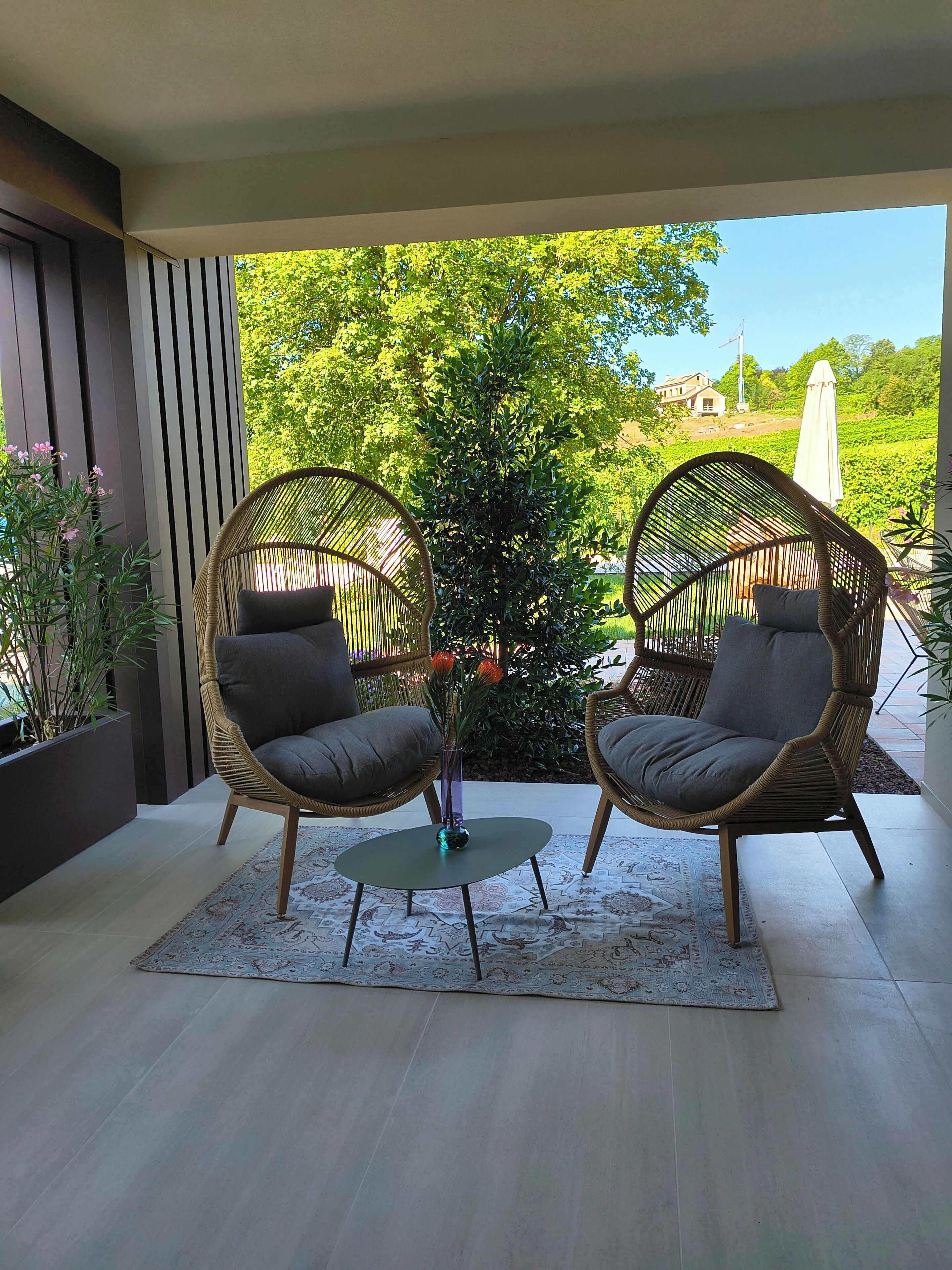 Ein überdachter Außenbereich mit Sitzgelegenheit, zwei Korbstühlen und einem kleinen Tisch, umgeben von einem Garten mit Blick auf ein Haus und Bäume.