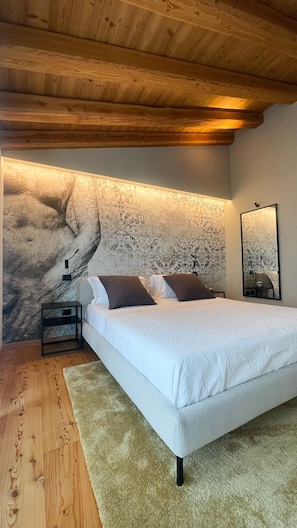 Un dormitorio con cama, suelo de madera, una mesita de noche, un espejo fijado a la pared y un papel tapiz estampado.
