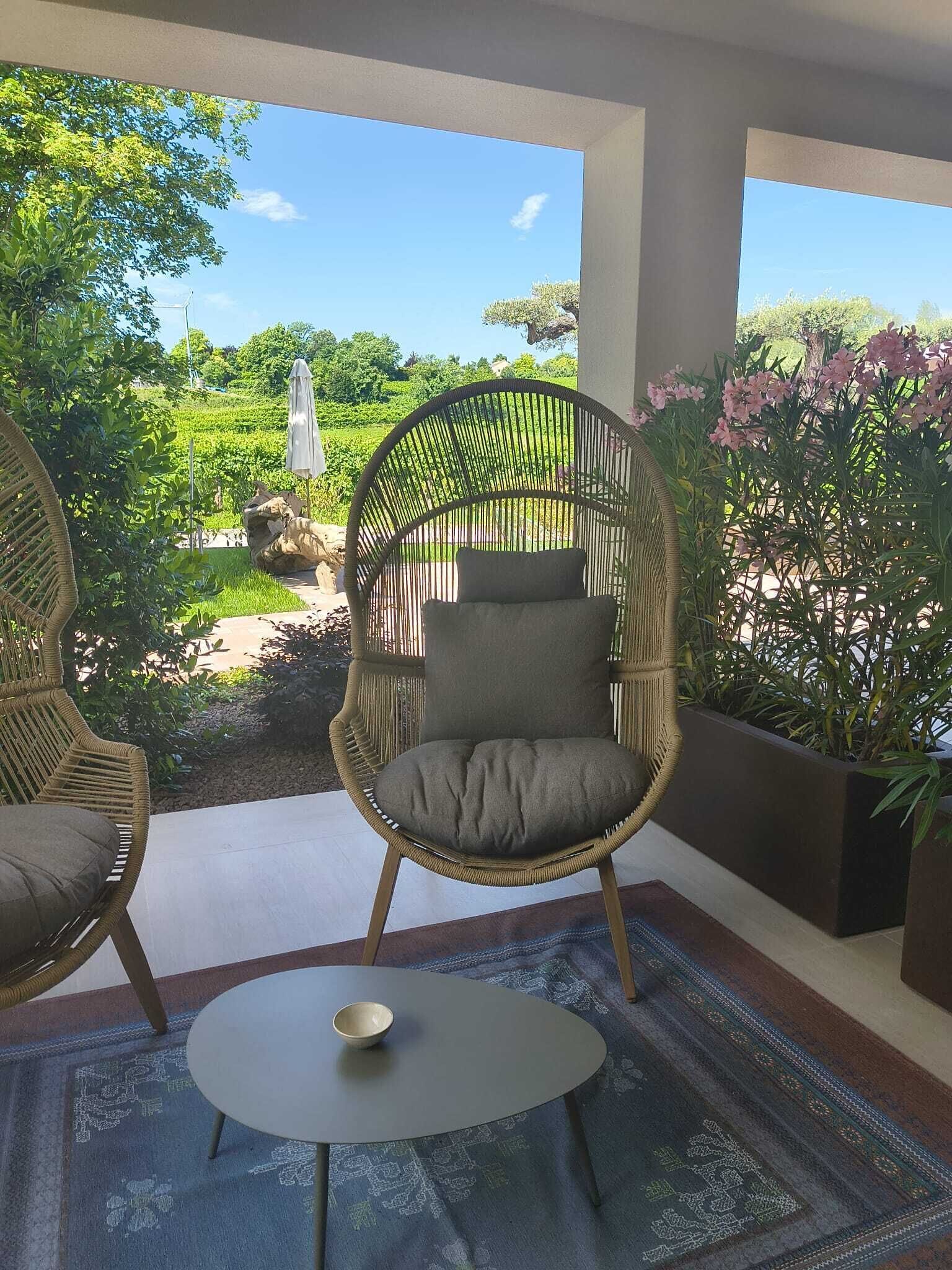 Una silla de mimbre con cojín, una mesita con un cuenco, y vista a un jardín con follaje y un molino.