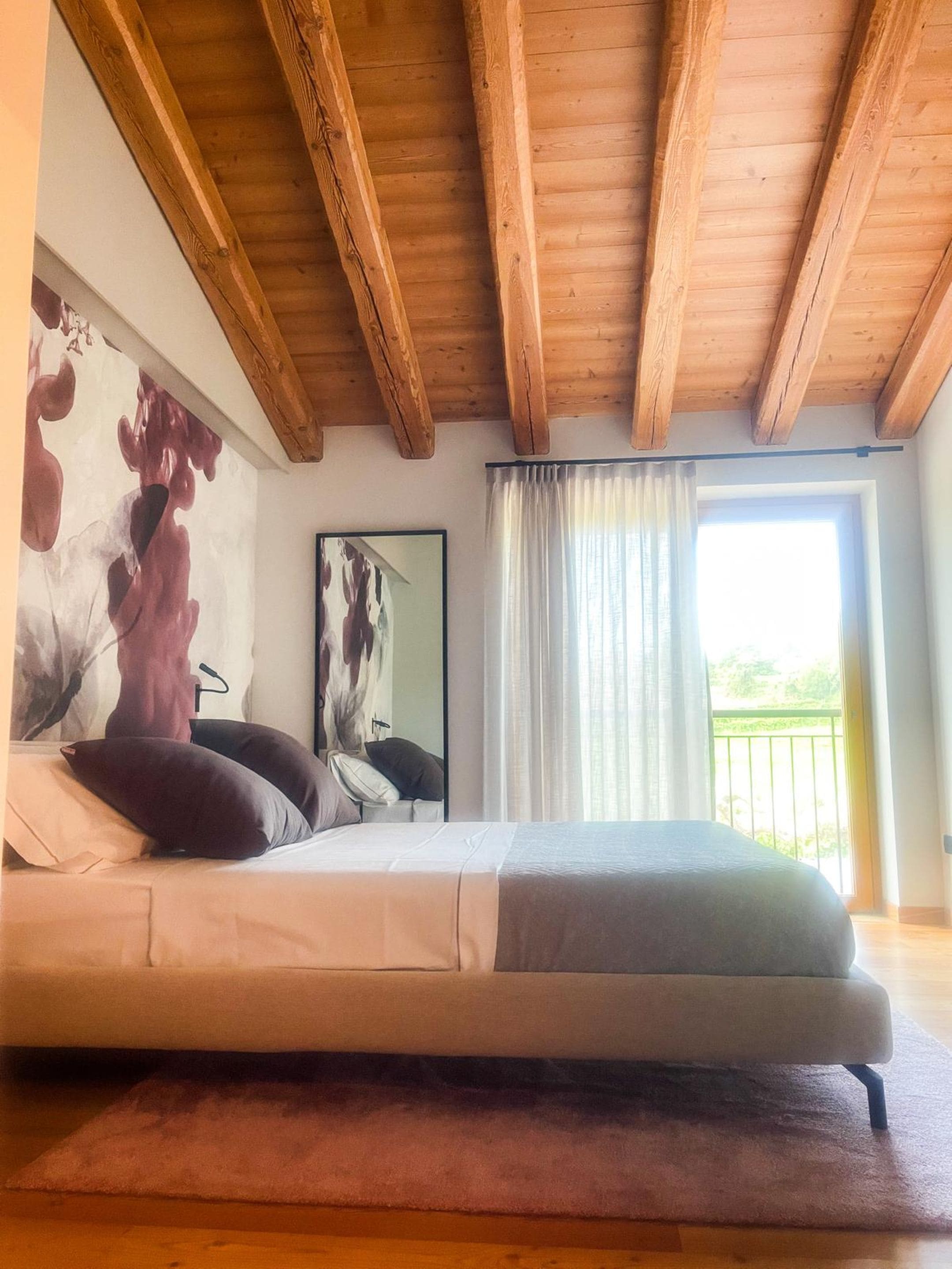 Ein Schlafzimmer mit Bett, Holzbalkendecke, einem großen Fenster mit Vorhängen und Blick auf Grün.
