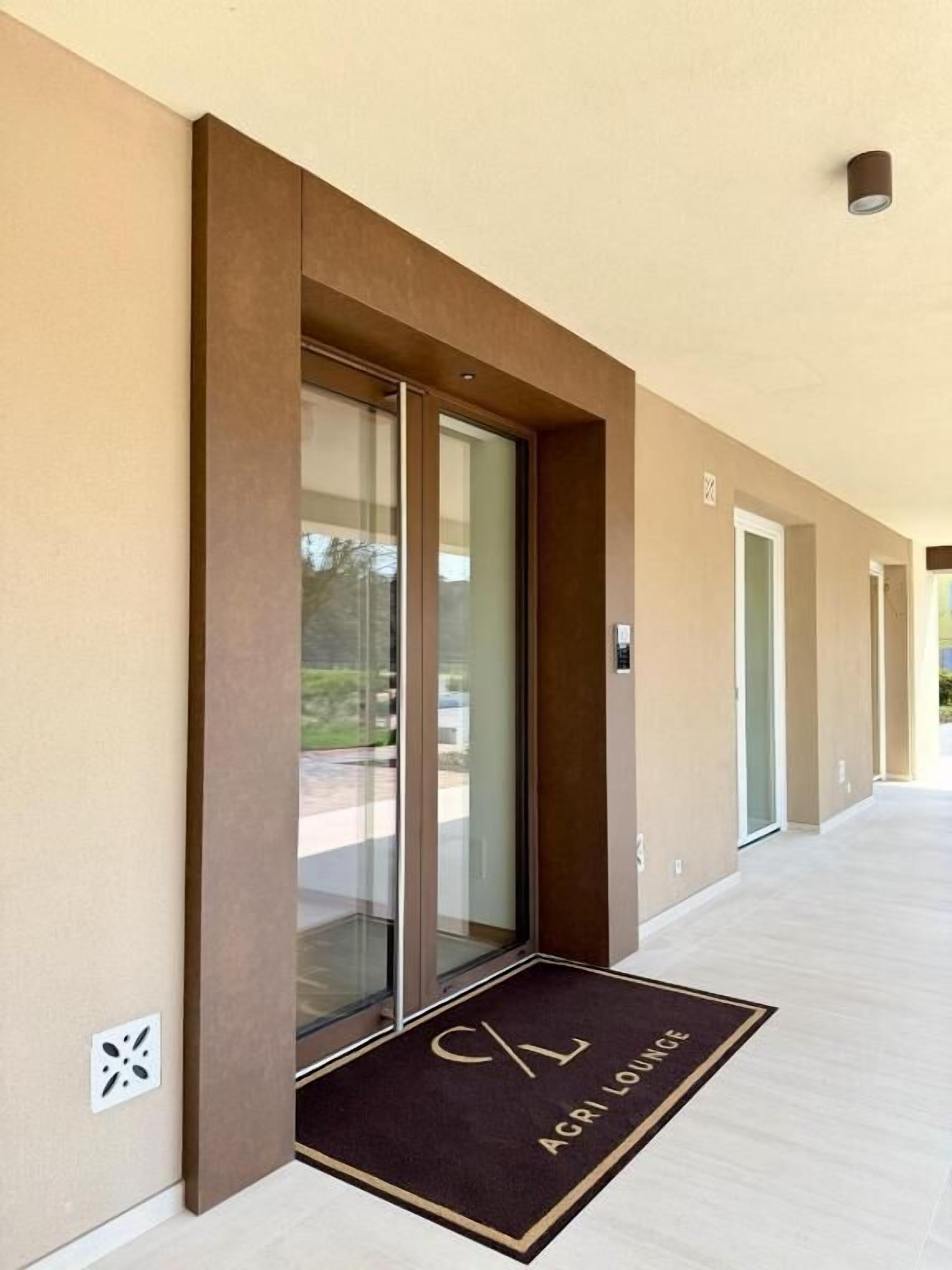 A modern building entrance with a glass door, a doormat with 'ACRI LOUNGE' text, and a light-colored corridor.