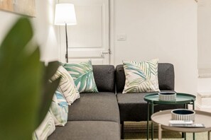 Una sala de estar moderna con un sofá gris, dos almohadones estampados, una lámpara de pie y dos mesitas redondas con adornos.