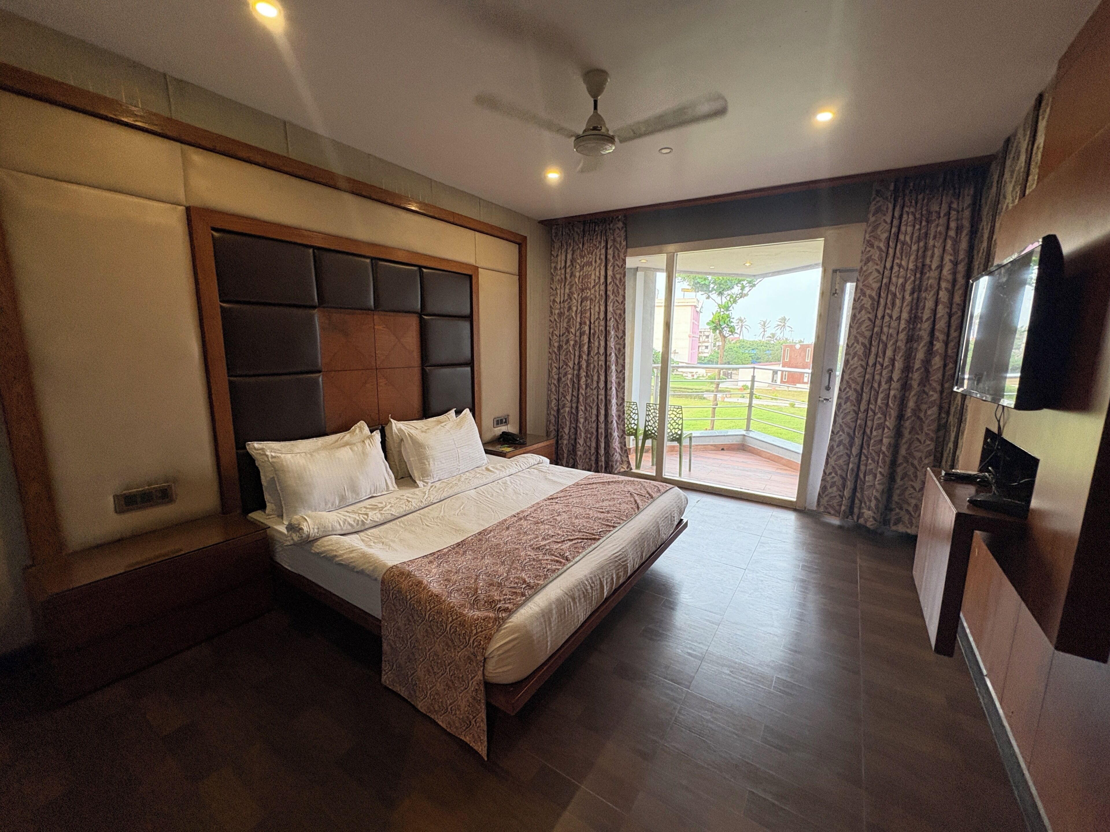 A bedroom with a large bed, a ceiling fan, a TV, and a view of the outside.