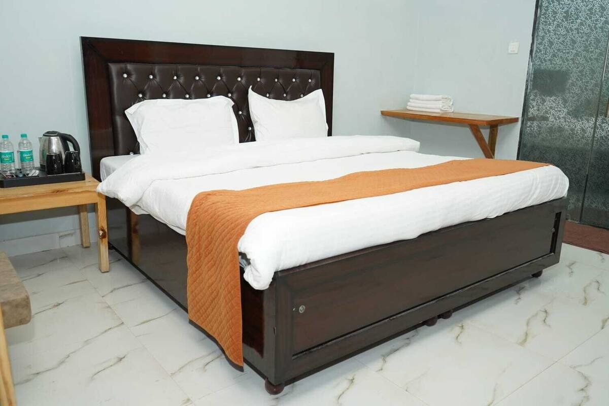 Una cama con cabecera oscura, ropa de cama blanca y una manta color naranja. Junto a la cama, una mesita de noche de madera con una tetera y botellas de agua.