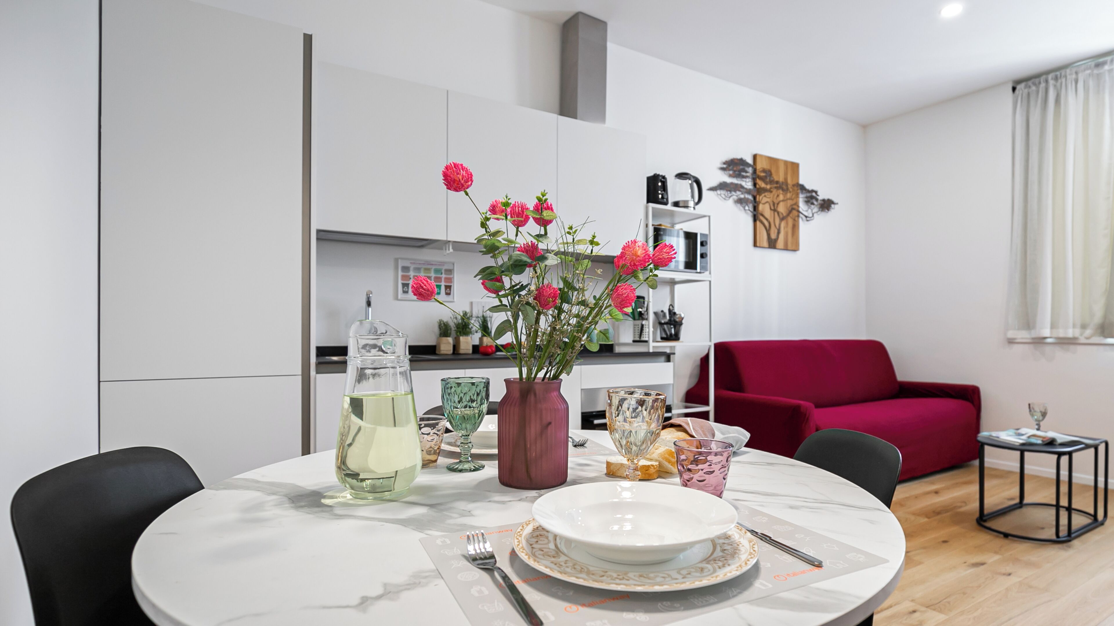 A modern living room with a dining table set for two, a red sofa, and a kitchen in the background.