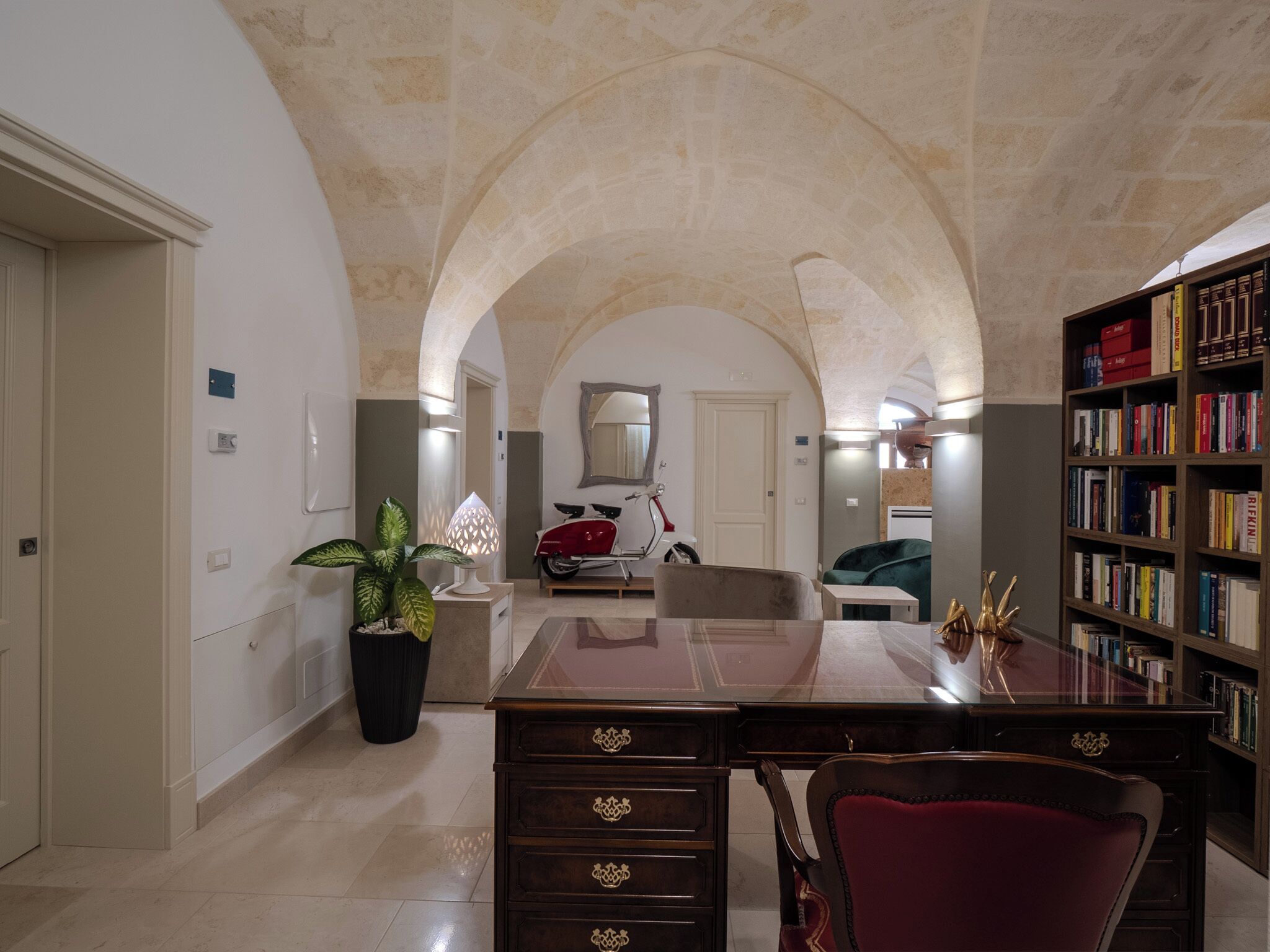 A spacious room with a high arched ceiling, a large desk with drawers, a bookshelf filled with books, and a potted plant.