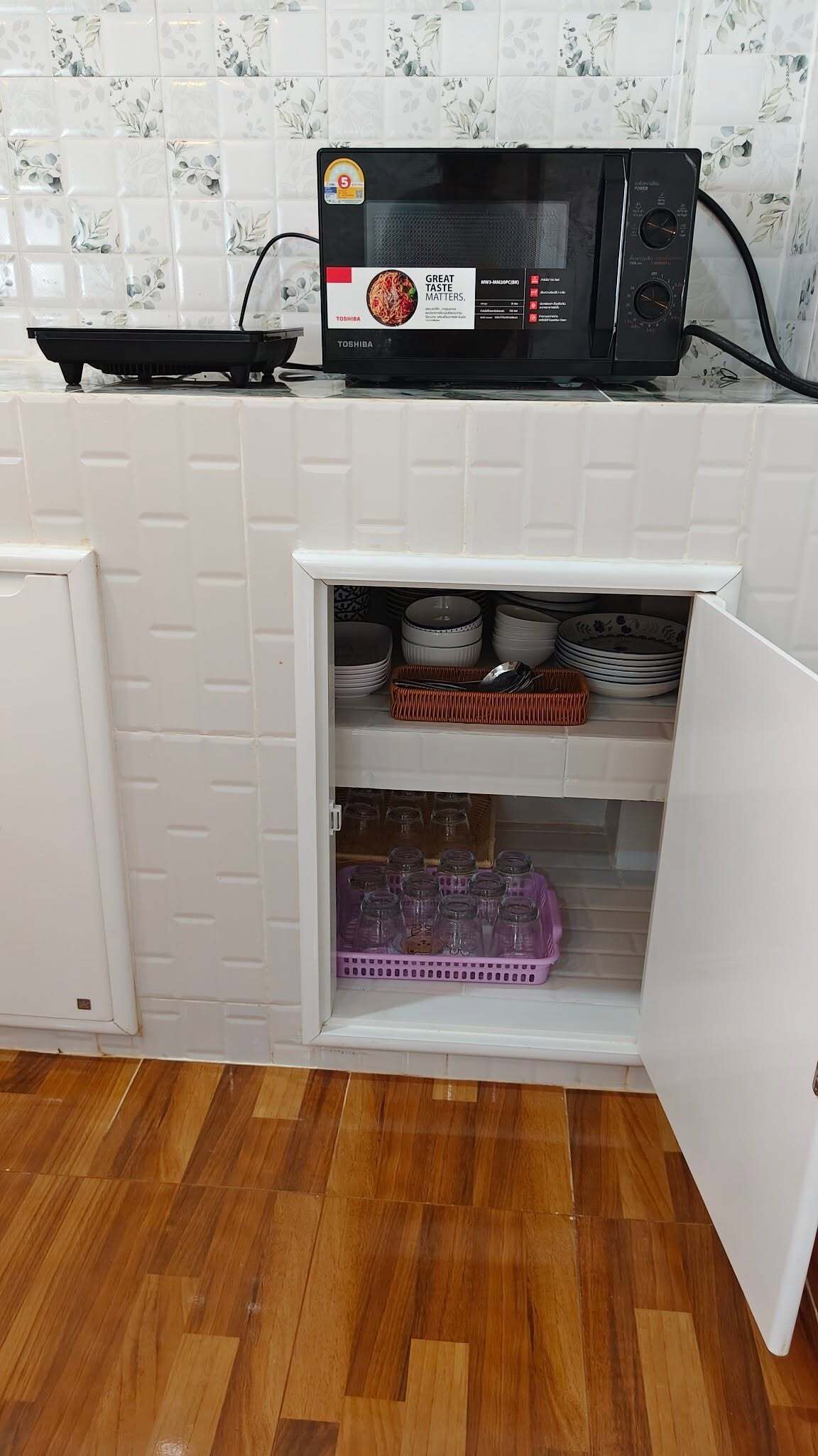 A compact kitchen with a microwave, a small refrigerator, and a storage cabinet containing dishes and jars.