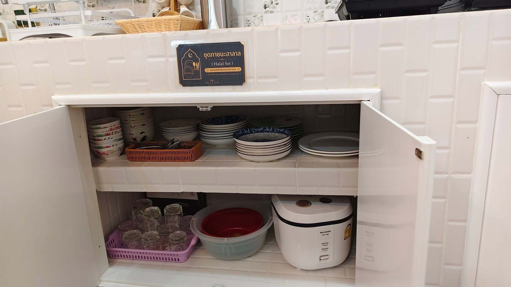 A kitchen cabinet with a halal set label, stacked plates, bowls, a rice cooker, and a purple glass container.