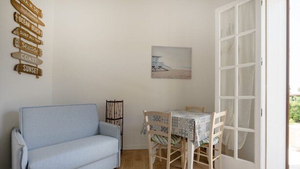 Quarto com um sofá azul claro, uma mesa de jantar com cadeiras, um quadro com uma cena de praia e uma parede com placas de madeira.