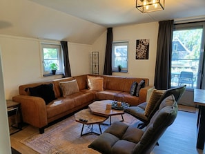 A living room with a brown leather sofa, a wooden coffee table, and a patterned rug. There are two windows with curtains, a potted plant, and a framed artwork on the wall. - Secret van Otterlo Luxe 10 (Otterlo)