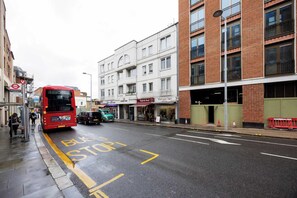 Ein roter Bus fährt auf einer Stadtstraße, an der ein Bushalteschild und ein Gebäude mit Geschäften zu sehen sind.
