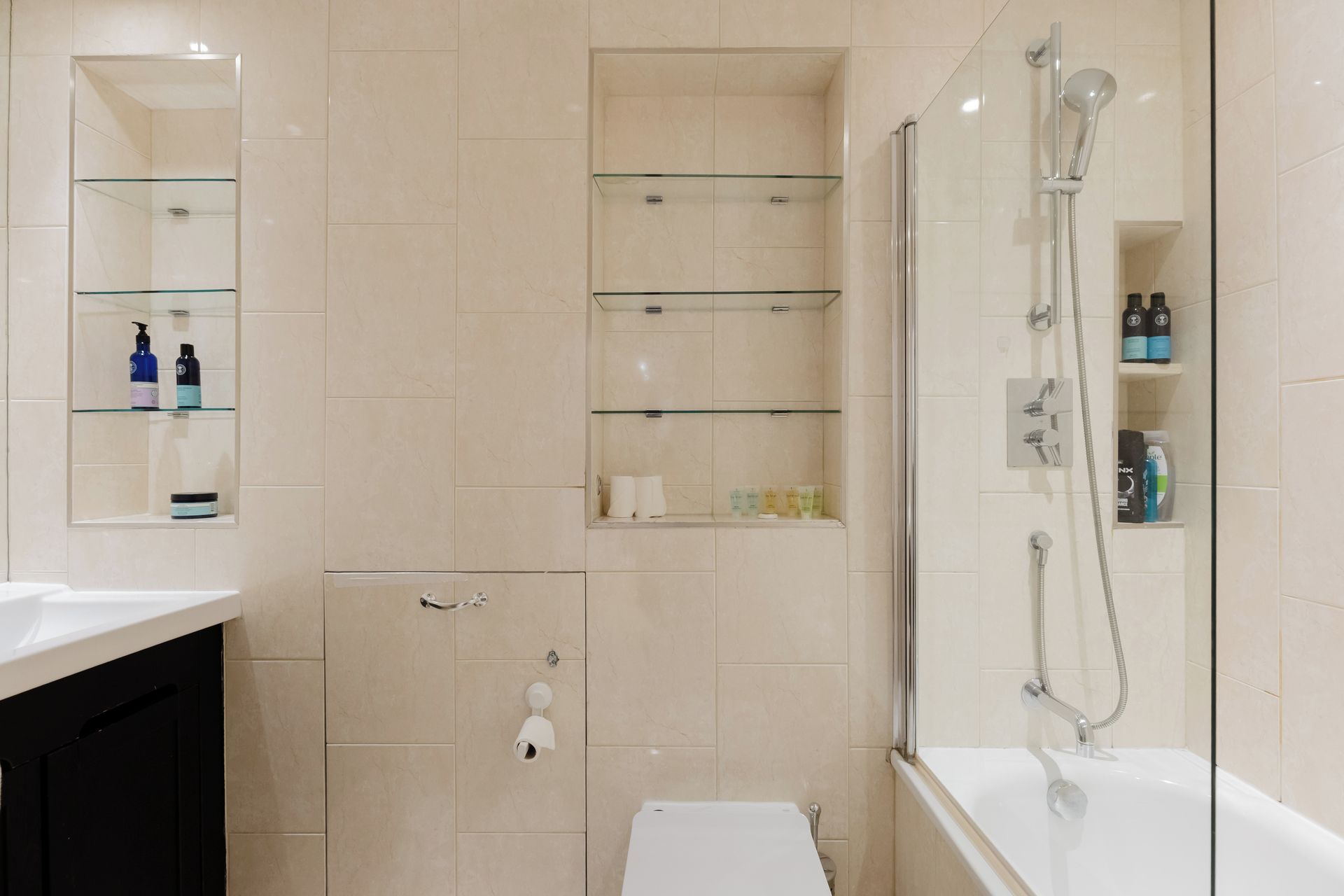 Une salle de bain moderne avec une douche à parois de verre, une baignoire blanche et des étagères en verre où sont rangés les produits de toilette.