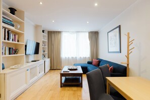 A modern living room with a flat-screen TV, a bookshelf, a blue sofa, and a wooden coffee table. - Inviting 1BDR Flat in Earls Court (Greater London)