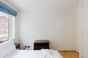 A bedroom with a bed, a wooden nightstand, a wall socket, and a window with a blue curtain. - Inviting 1BDR Flat in Earls Court (Greater London)