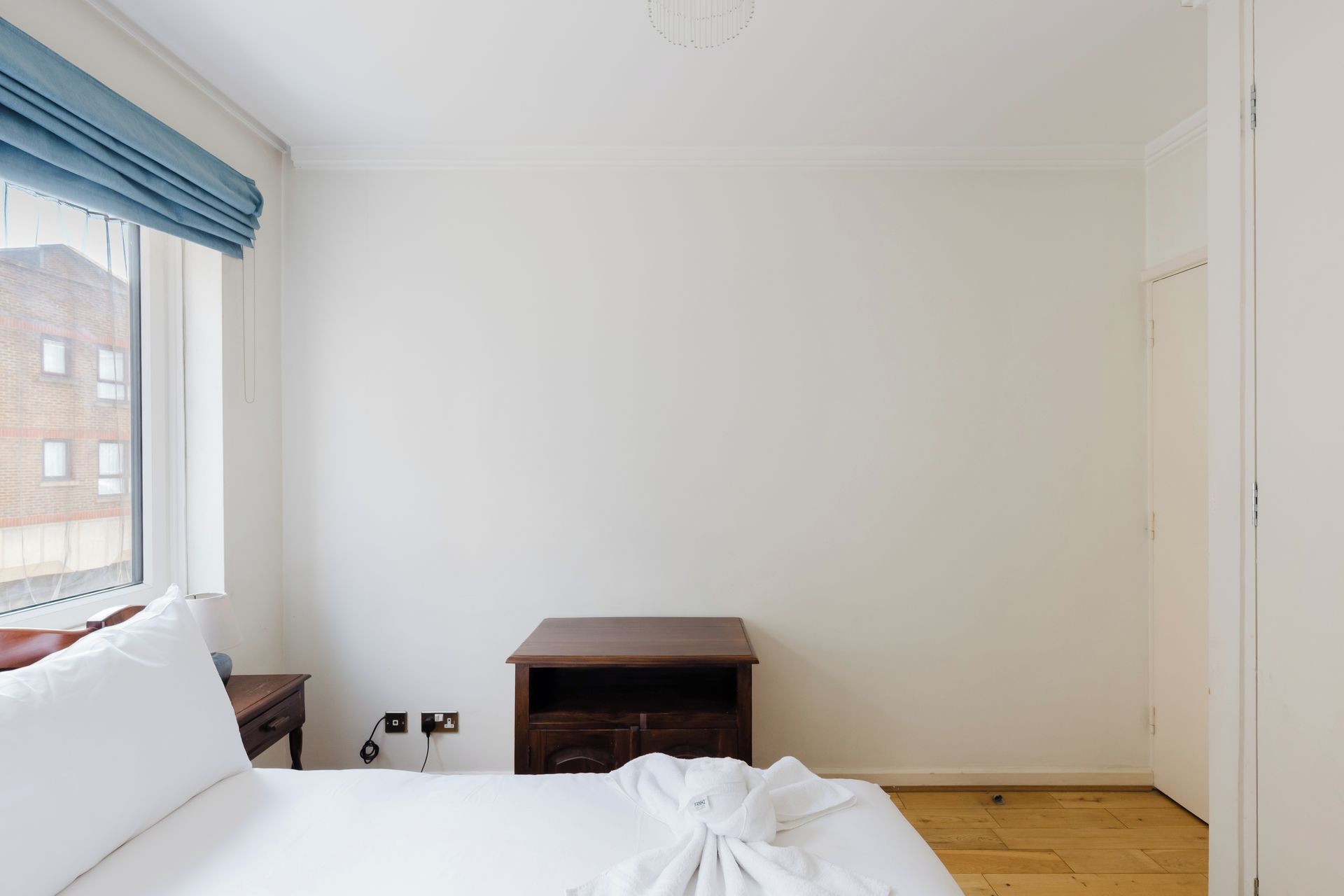 A bedroom with a bed, a wooden nightstand, a wall socket, and a window with a blue curtain.