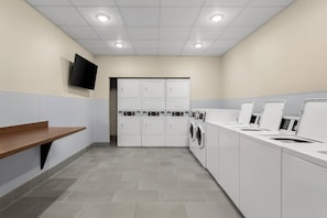 A laundry room with multiple washing machines, a flat-screen TV mounted on the wall, and a wooden bench.