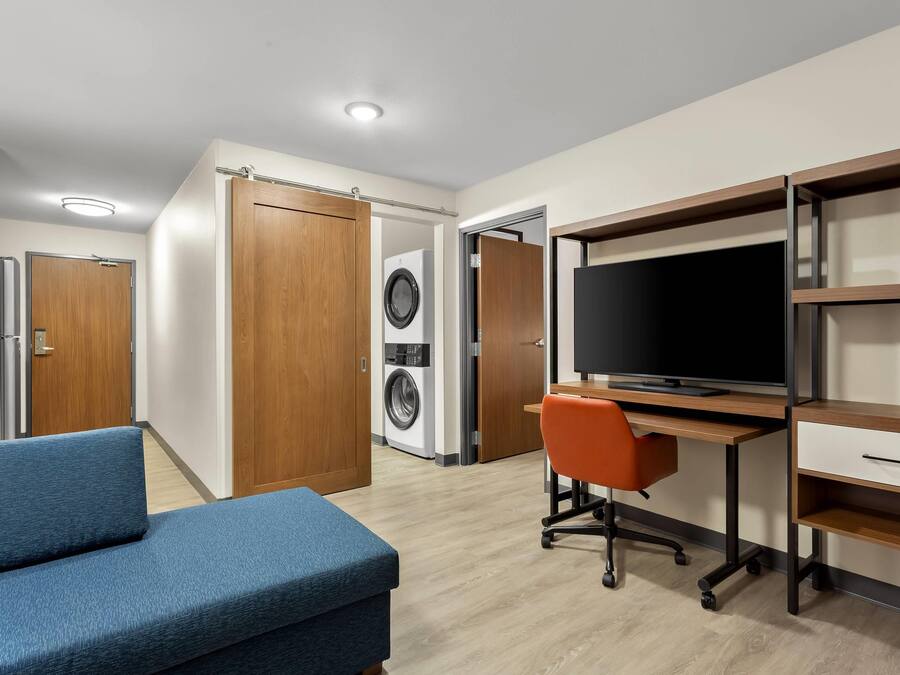 A modern bedroom with a bed, a desk with a computer, a TV, and a washing machine.