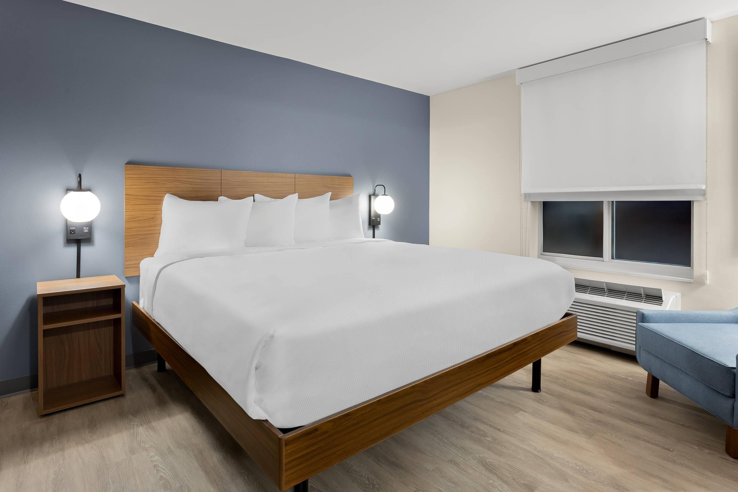 Un dormitorio moderno con estructura de cama de madera, ropa de cama blanca, dos lámparas de noche, una mesita de noche, una silla azul y una ventana con persianas.