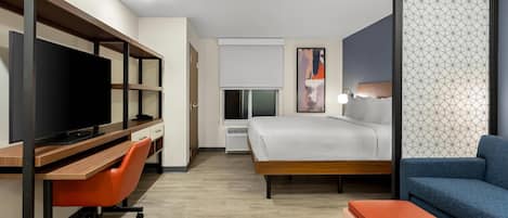 Habitación de hotel con una cama, un escritorio con televisor, una silla, un sofá y una pared divisoria con un diseño geométrico.