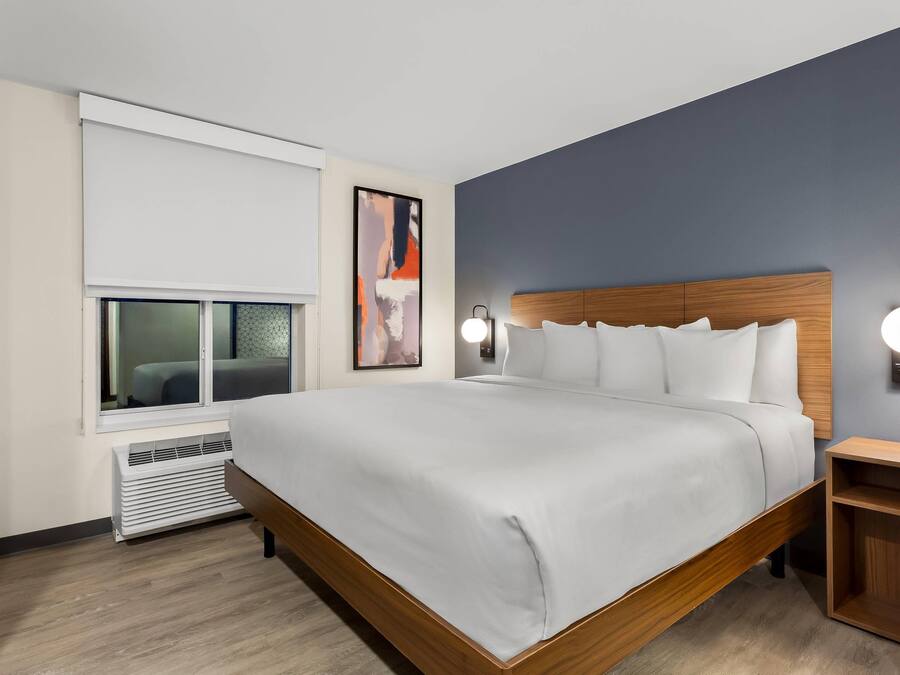A modern hotel room with a large bed, a TV, a wall-mounted artwork, and a bedside table with a lamp.