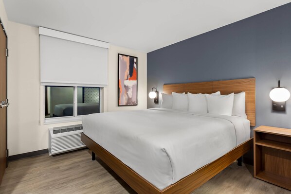A modern hotel room with a large bed, a TV, a wall-mounted artwork, and a bedside table with a lamp.