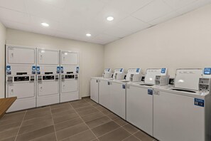 Een wasserette met meerdere wasmachines en drogers, een betegelde vloer en een wit plafond met inbouwverlichting.