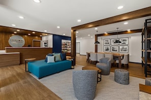Un moderno vestíbulo de hotel con mostrador de recepción, un sofá azul, sillones grises, una zona de comedor y cuadros en la pared.