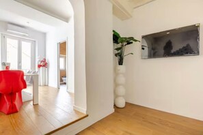 Un corridoio con pavimento in legno, una sedia rossa, un vaso bianco con piante e un quadro appeso al muro.