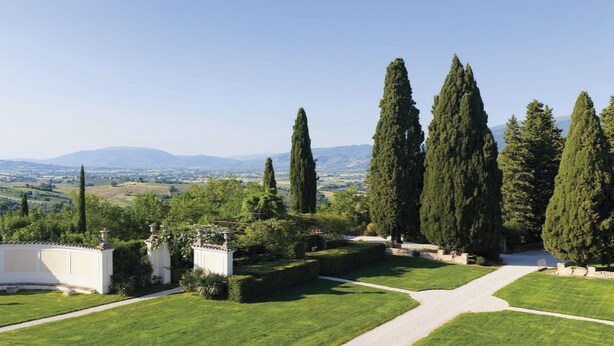 A landscaped garden with a gravel path, tall cypress trees, and a view of distant hills.