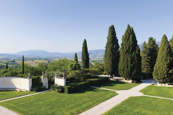 Un jardin aménagé avec un chemin de gravier, de grands cyprès et une vue sur des collines lointaines.
