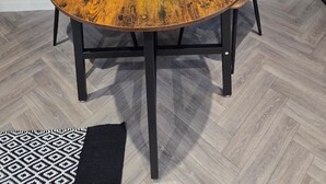 Een ronde houten tafel met een witte placemat en een gele bloemdecoratie, omringd door twee bruine fauteuils, voor een donkere wand met verticale panelen.