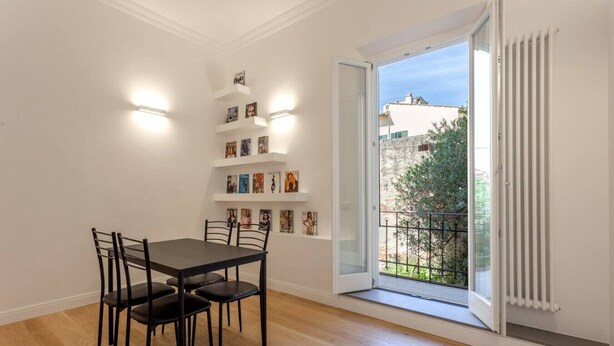 Zona pranzo con tavolo e sedie, una libreria piena di libri, una finestra con vista su edifici e vegetazione, e un termosifone.