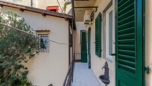 Un stretto corridoio esterno con pavimento piastrellato, persiane verdi e una piccola finestra con sbarre di metallo.