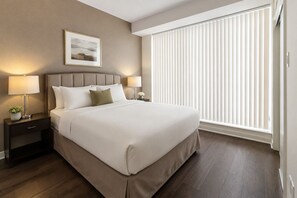 A hotel room with a large bed, two bedside tables with lamps, a framed picture on the wall, and a window with blinds. - Merona Grand Hotel (Toronto)