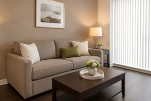 A living room with a beige sofa, a dark coffee table, a lamp, and a framed picture on the wall. - Merona Grand Hotel (Toronto)