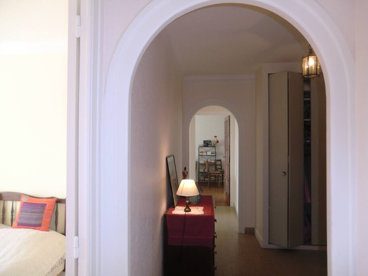 Un pasillo estrecho con un arco que da a un dormitorio, con una cama y una mesita de noche con una lámpara.