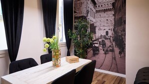 Una habitación con una mesa, sillas, un florero con flores, una caja de bienvenida y un mural que representa una escena de calle histórica.