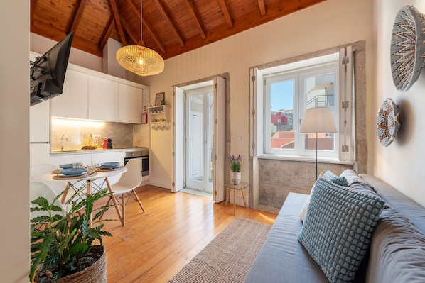 Ein modernes Interieur mit Holzbalkendecke, einem Essbereich mit Tisch und Stühlen, einer Couch mit gemusterten Kissen und Blick durch eine offene Tür.