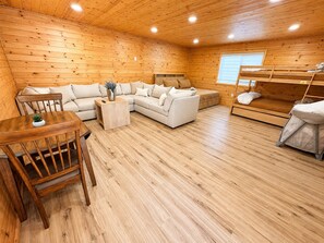 Ein gemütliches Blockhäuschen-Interieur mit Holzboden, einer Sofagarnitur, einem Esstisch mit Stühlen und einem Etagenbett.