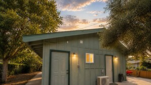 A small, single-story building with a green exterior, a front door, two windows, and an air conditioning unit on the ground. - Rock House/Riverfront/EV Charging/Large Groups (Kernville)