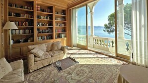 Uma sala de estar ampla com uma grande estante de livros, um sofá confortável, uma mesa de centro e vista para o mar através de grandes janelas.